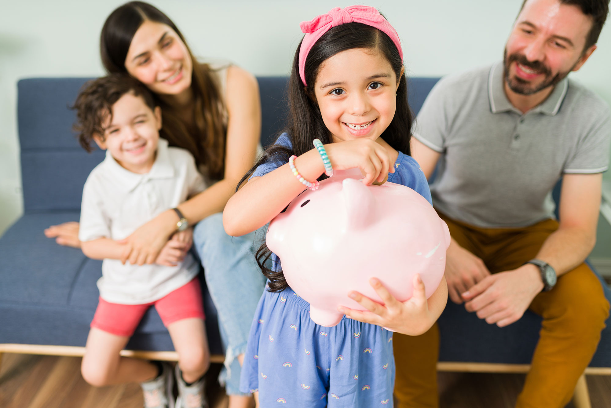 Young girl holding piggy bank while other families sit on couch behind her.