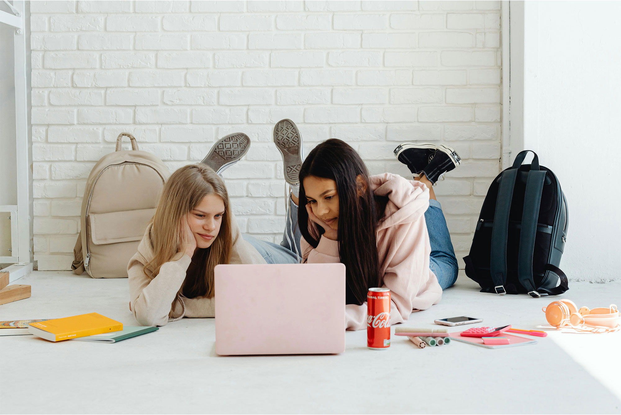 Two teen girls laying on floor with school supplies and bookbags near them looking together at laptop screen.